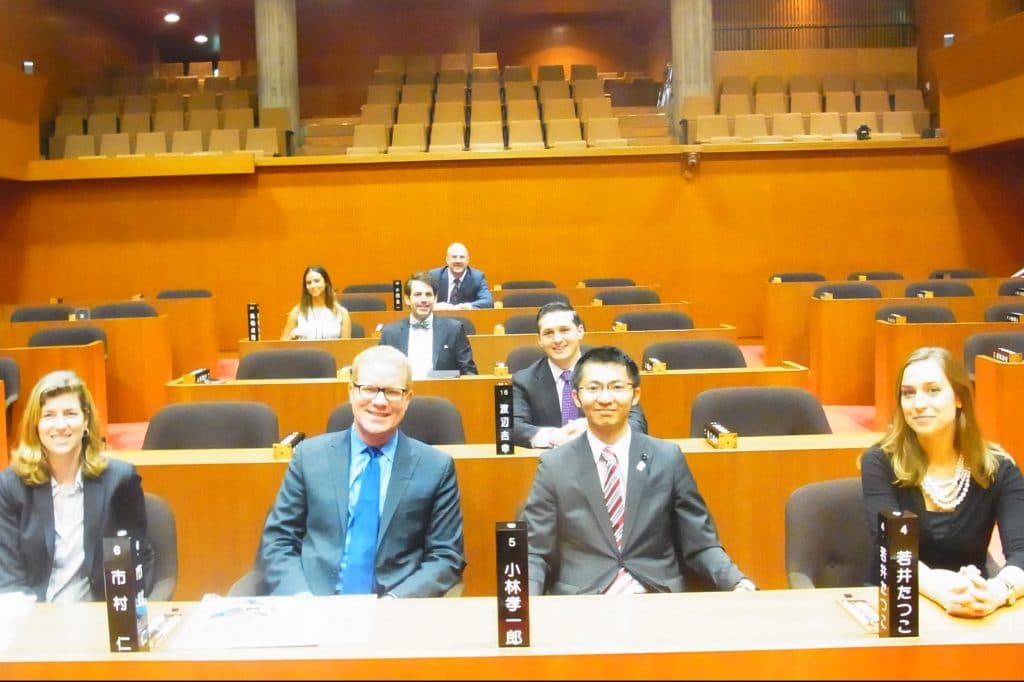 Delegation members try out the seats in the Okayama Prefectural Assembly chambers with Assembly Member Koichiro Kobayashi.